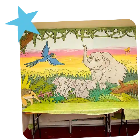 A colourful mural of a jungle scene brightens the space at Navan Adventure Centre, featuring three elephants, a blue parrot in flight, a butterfly, lush green foliage, and a vibrant sunset sky. A table covered with a green cloth sits in front of the mural.