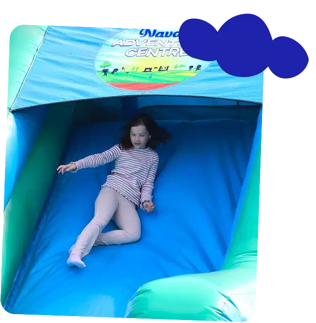 A young girl wearing a striped shirt and light trousers slides down a blue and green inflatable slide at the outdoor Navan Adventure Centre.