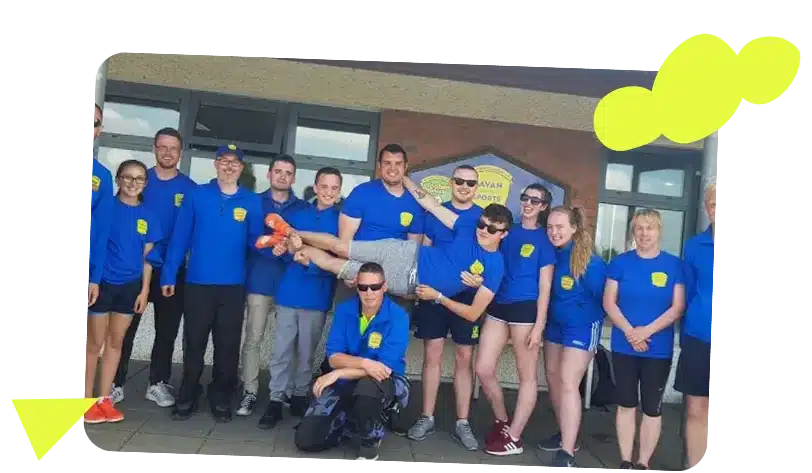 A group of people in matching blue shirts poses outside Navan Adventure Centre. One person is held horizontally by others at the centre, while the group stands smiling around them. Yellow graphic shapes decorate the image corners.