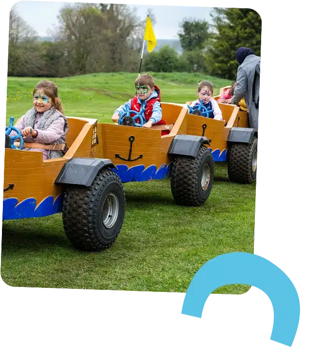 Three young children with face paint ride in a wooden cart train on grass at Navan Adventure Centre, each holding a steering wheel. An adult stands beside them, trees are visible in the background, and a yellow flag is attached to the first cart.