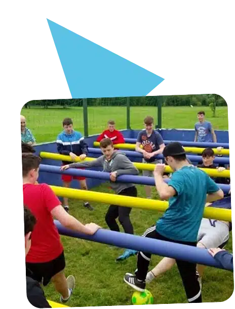 A group of teenagers are playing a life-sized human table football game outdoors at Navan Adventure Centre, each holding onto yellow and blue horizontal poles, with a green and yellow football on the grass.