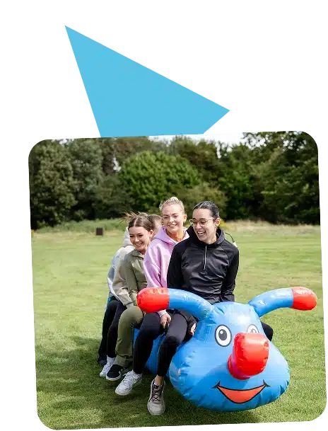 Three smiling women sit outdoors on a large blue inflatable toy shaped like an animal with red handles and a face at Navan Adventure Centre, whilst another woman stands behind them. Green grass and trees create a lively background.