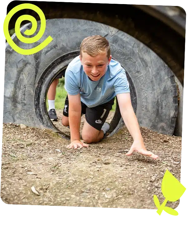 A smiling boy in a blue shirt crawls out of a large rubber tyre on a dirt playground at Navan Adventure Centre, with another child partially visible inside. Yellow spiral and spade icons decorate the image corners.