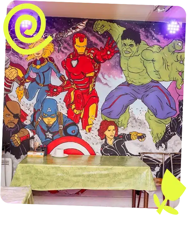 A wall mural of Marvel superheroes—including Captain Marvel, Iron Man, Hulk, Nick Fury, Captain America, and Black Widow—decorates a dining area at Navan Adventure Centre with a table, green tablecloth, and chairs.
