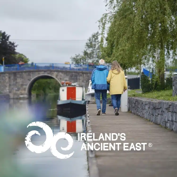 An older couple wearing blue and yellow jackets walk hand in hand along a canal towpath near Navan, with a moored boat and stone bridge in the background. Text reads "Ireland's Ancient East" with a white logo, highlighting things to do in Navan.