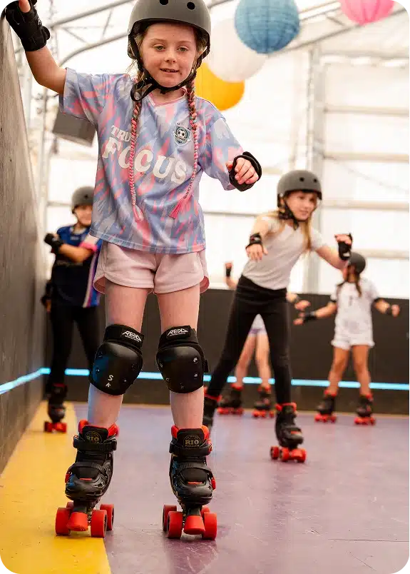 A young girl wearing a helmet and knee pads roller skates on an indoor rink, smiling at the camera. Other children on skates are skating behind her. Colourful lanterns hang above.