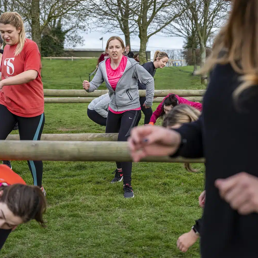 A group of women exercise outdoors on a grassy field at the Navan outdoor adventure obstacle course, climbing under and over wooden barriers in sportswear. Trees and cloudy skies form the scenic background as they focus on the activity.