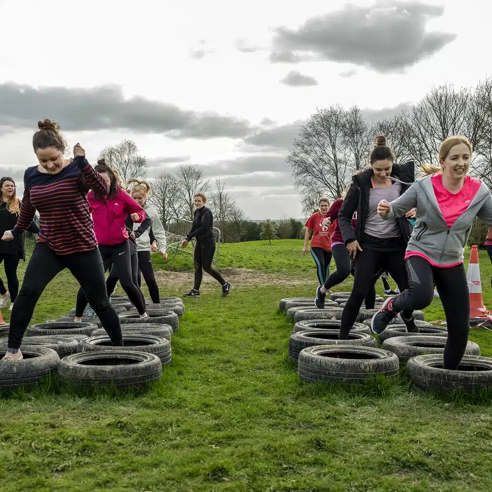 A group of women tackles the Navan outdoor adventure obstacle course, stepping through tyres on a grassy field under a cloudy sky. Some are smiling as they run, with trees visible in the background.