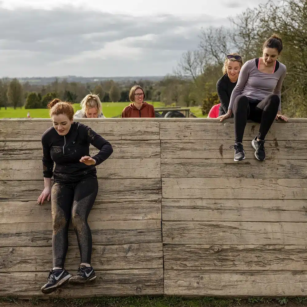 Four women are at the Navan outdoor adventure obstacle course. Two are climbing over a wooden wall, whilst two others cheer them on in the background. The scene is set in a grassy area with trees and cloudy skies.