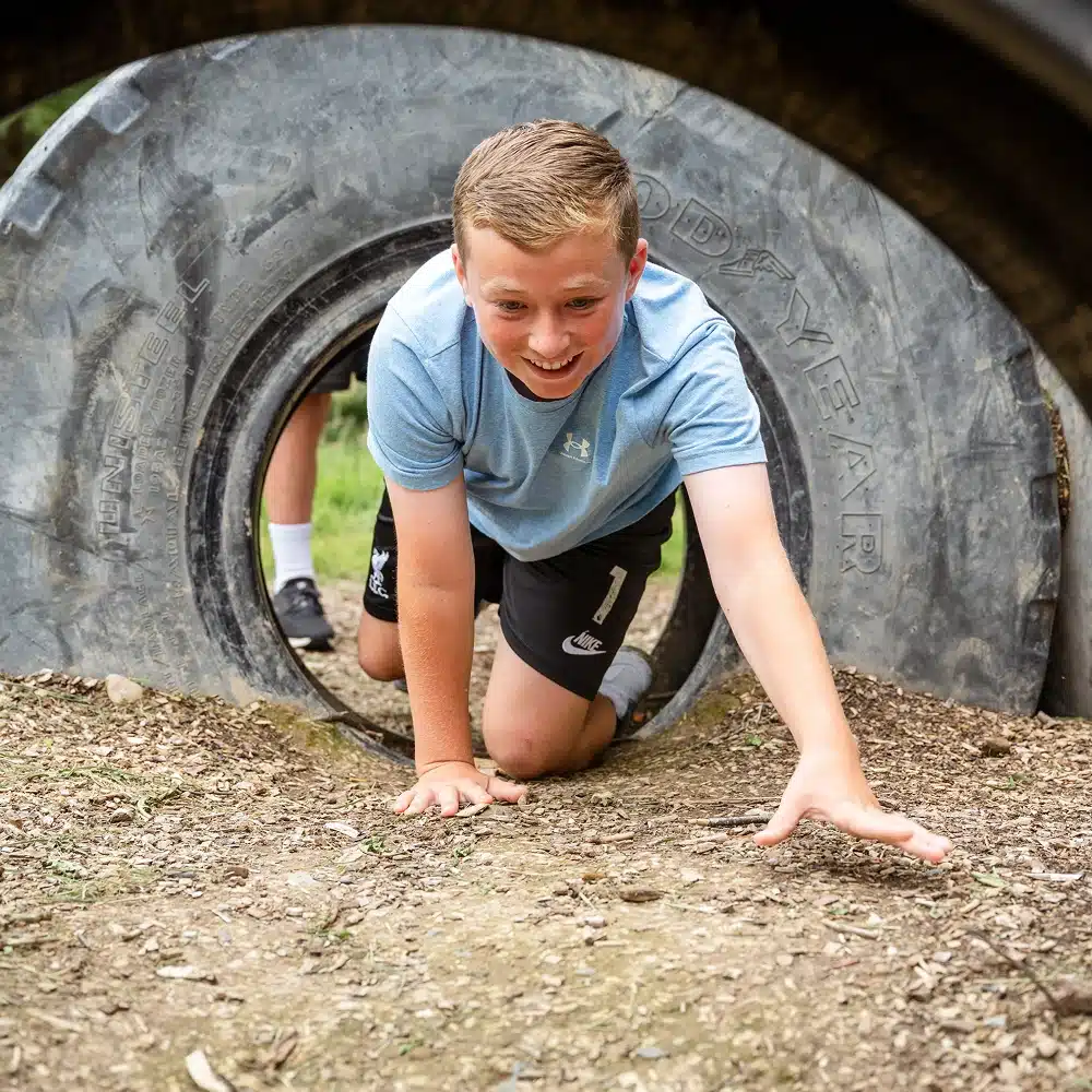 A smiling boy in a light blue shirt and black shorts crawls through a large tyre on the Navan outdoor adventure assault course, reaching forward with one hand. Another person’s legs are visible behind him.
