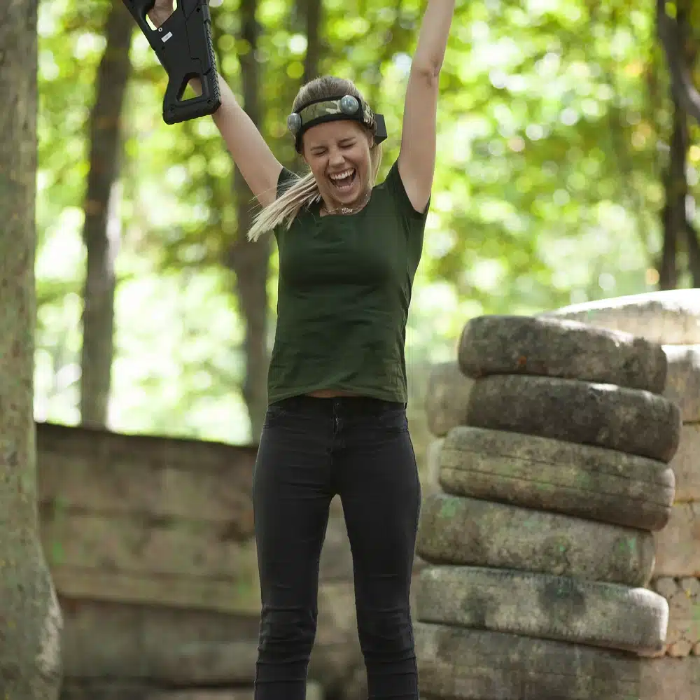 A woman outdoors raises her arms and smiles triumphantly while wearing a headband and holding a laser tag gun, enjoying Navan team building activities amid trees and stone barriers in the background.