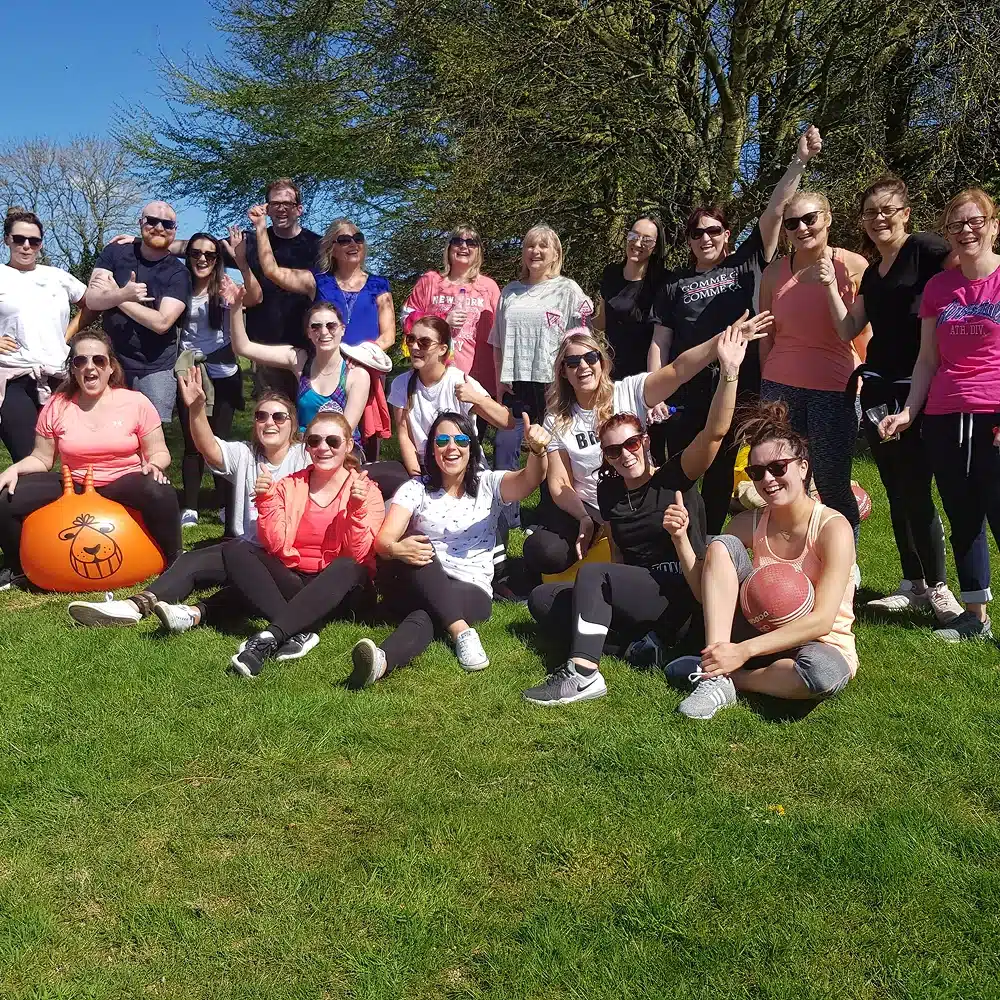 A group of smiling people pose together outdoors on a sunny day, sitting and standing on green grass with trees in the background. Some are wearing sunglasses and sporty clothing, enjoying Navan team-building activities with sports equipment in hand.