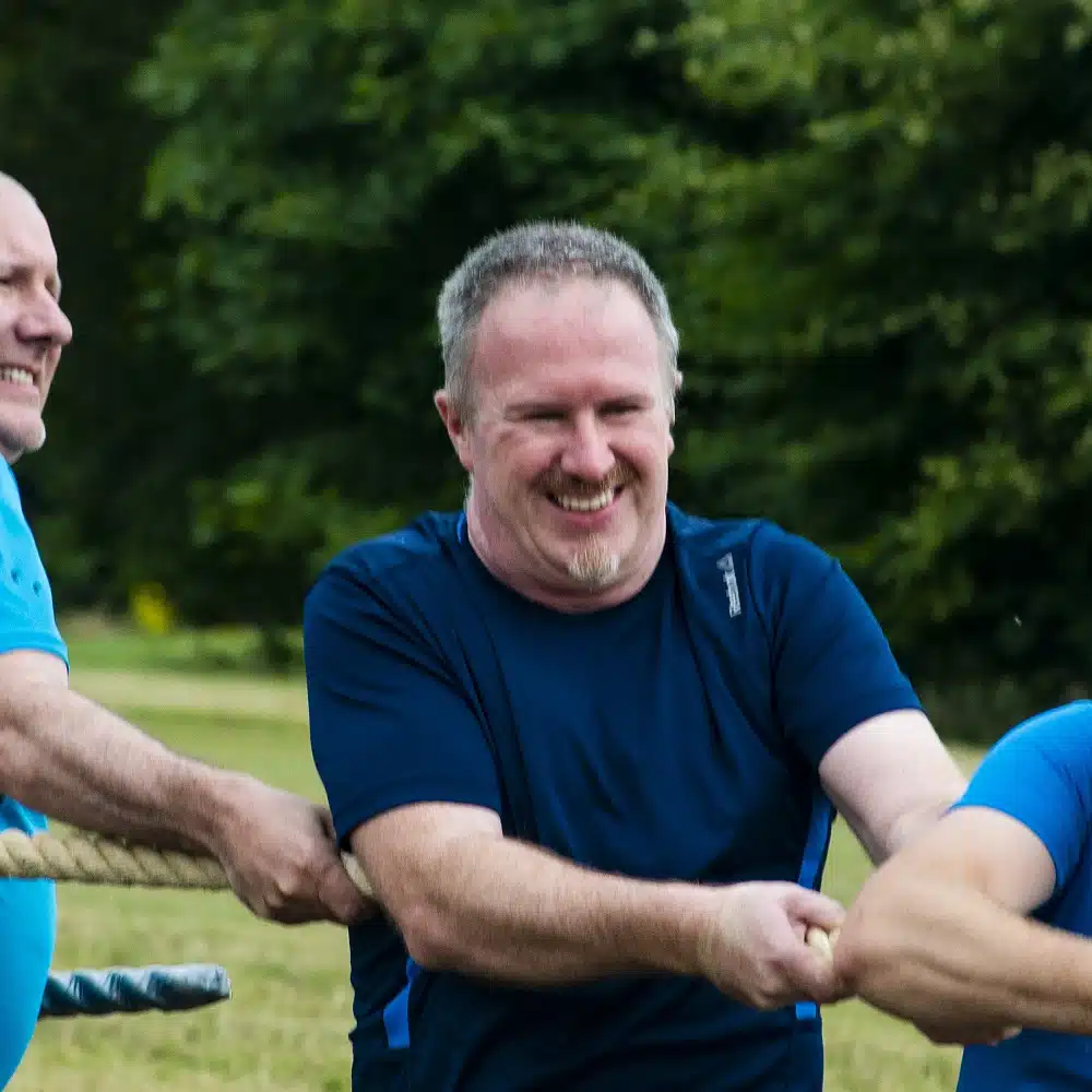 A smiling man in a blue shirt is pulling on a rope during an outdoor tug-of-war game—a perfect example of Navan team-building activities—with greenery and other participants visible in the background.