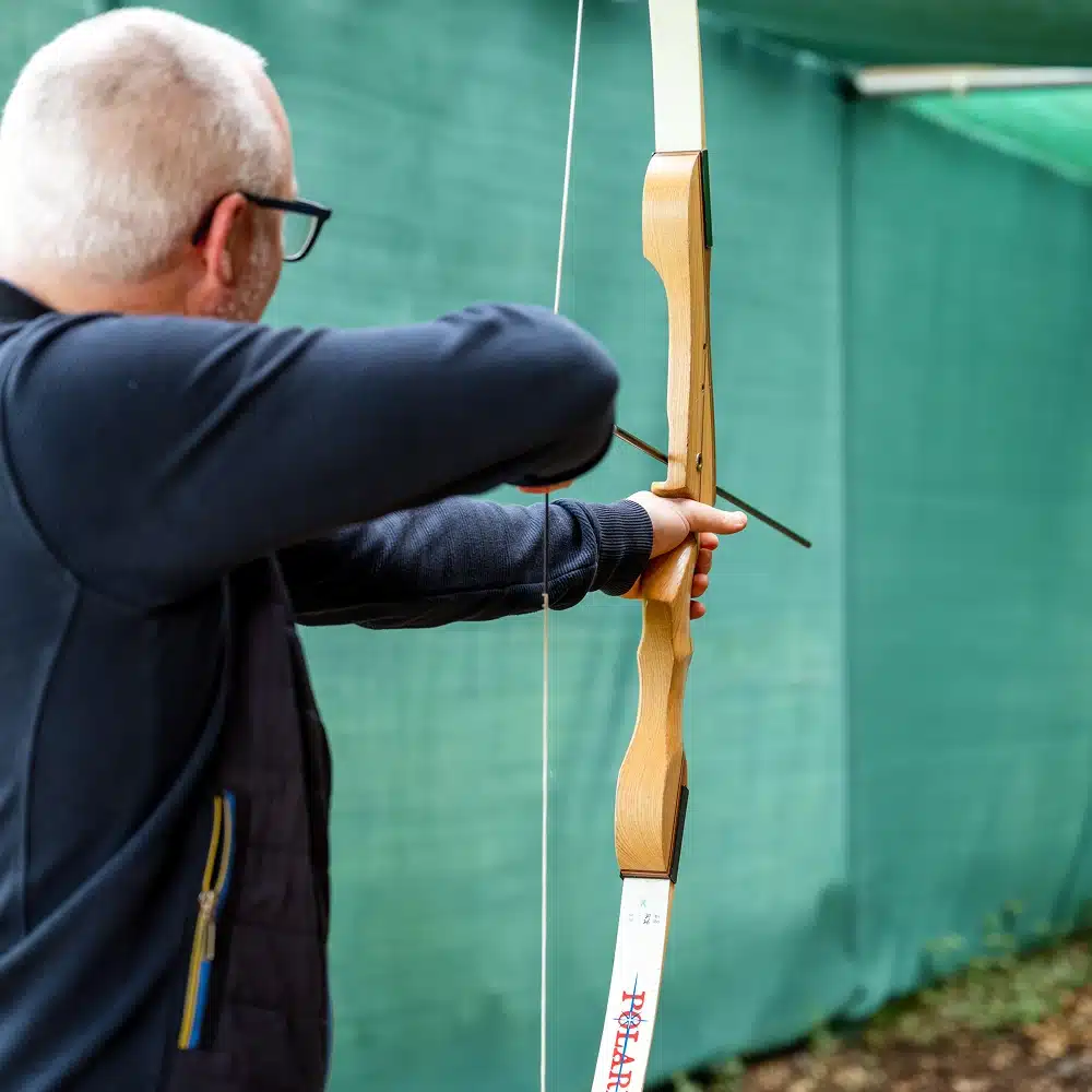 A person with short grey hair and glasses aims a wooden recurve bow, drawing an arrow back whilst standing near a green fabric background outdoors, practising skills honed at Navan indoor archery.