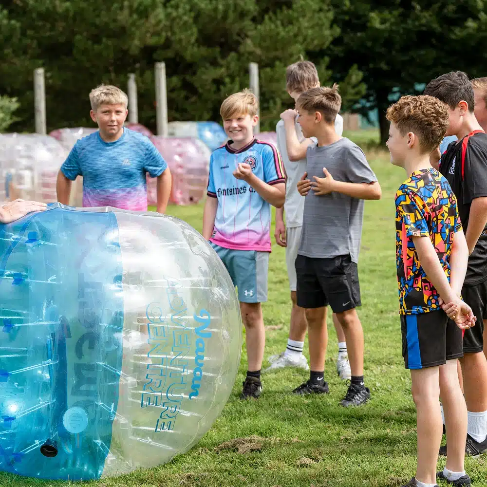 A group of boys stand on grass outdoors, smiling and laughing near a large blue and clear inflatable bubble-ball at a Navan bubble football event, with trees and more bubble-balls in the background.