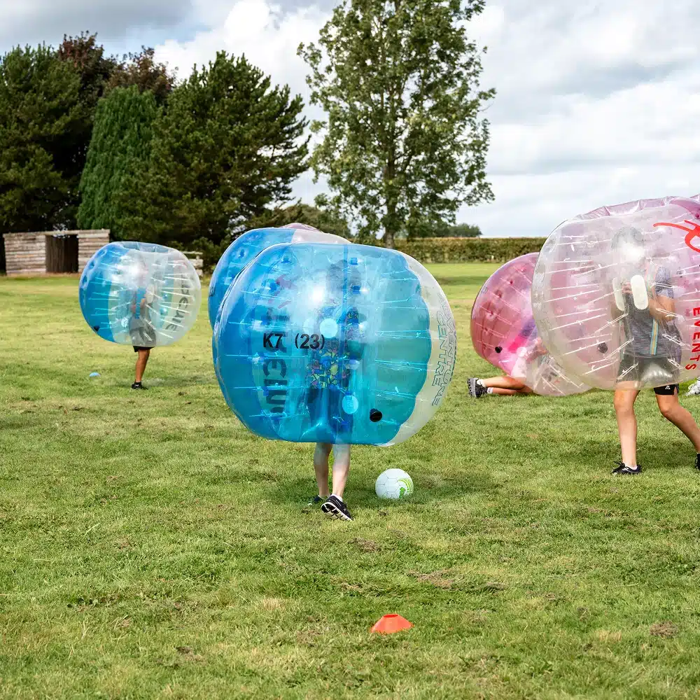 Three people wearing large inflatable bubble suits play Navan bubble football on a grassy field. Trees and a small building are in the background, cones mark the play area, and a blue and white football is near one player.
