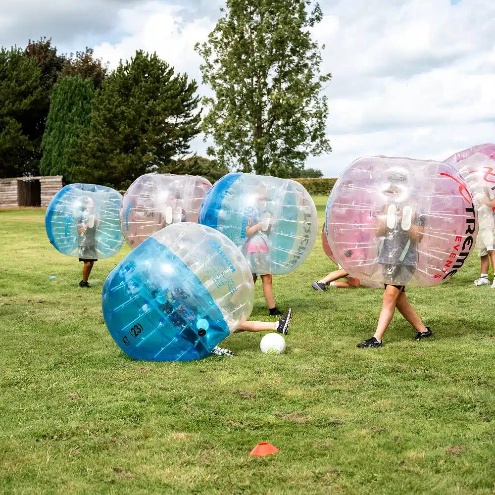 Children play football outdoors in Navan bubble football suits, with one child falling over on the grass. Trees and a cloudy sky are visible in the background.