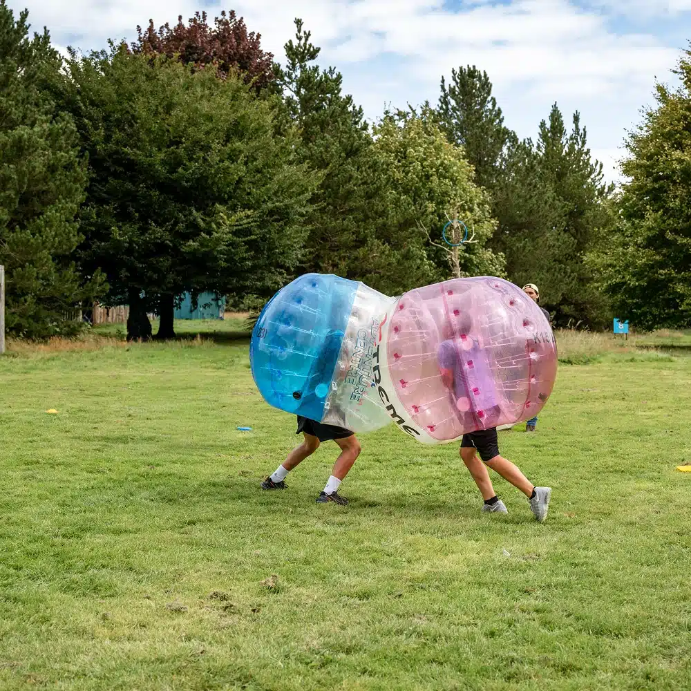 Two people wearing large inflatable bubble suits, one blue and one pink, collide on a grassy field surrounded by trees—an action-packed moment from Navan bubble football, with only their legs visible outside the bubbles.