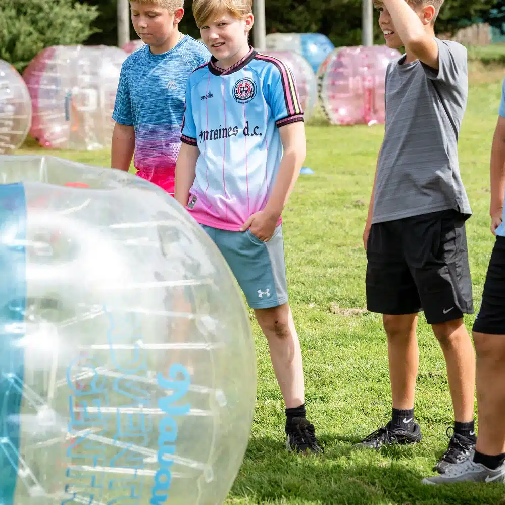 Three boys stand on grass next to a large, clear inflatable bubble ball, with more bubble balls in the background. They appear to be outdoors on a sunny day, possibly getting ready for a Navan bubble football match.