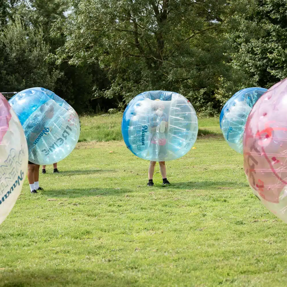 Several people are enjoying Navan bubble football outdoors, each inside a large inflatable bubble suit on a grassy field with trees in the background.