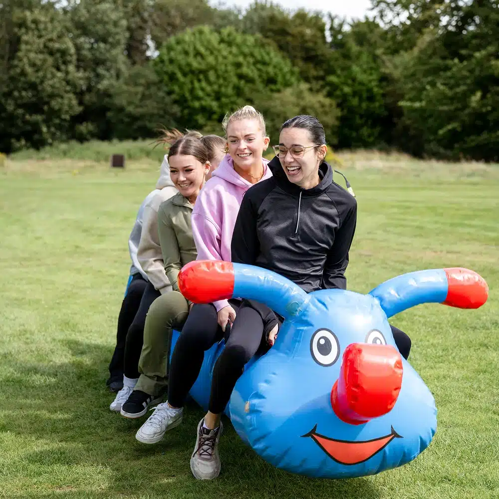 Four women sit in a row on a large, blue inflatable toy with a smiling face and red handles, outdoors on a grassy field. Laughing together, they enjoy one of the exciting Navan team building activities surrounded by trees.