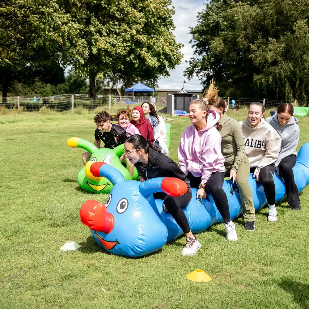A group of people laughing and riding inflatable caterpillar toys on a grassy field during Navan team building activities, with trees and tents visible in the background.