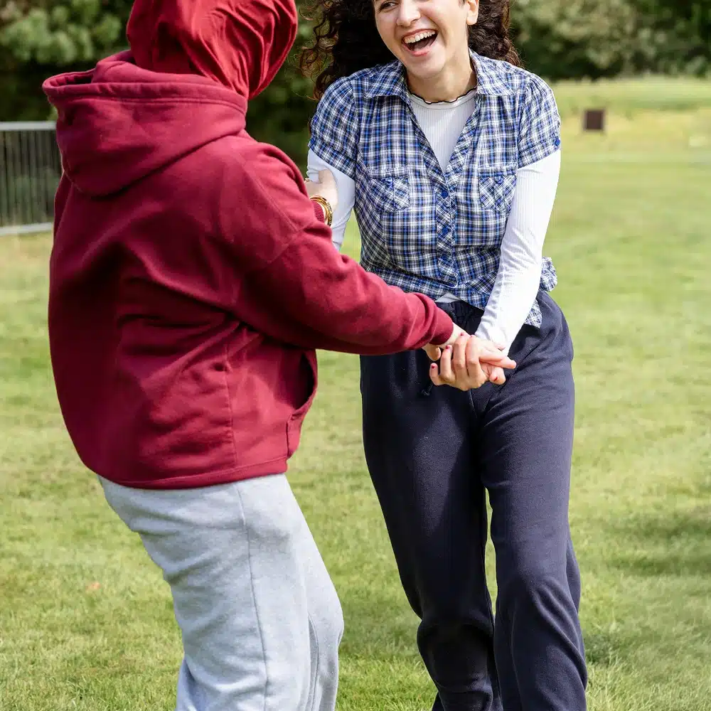Two people holding hands and smiling whilst playing outside on green grass, enjoying Navan team-building activities. One wears a red hoodie and grey trousers; the other sports a blue checked shirt, white top, and navy trousers. Trees and a fence are visible behind them.