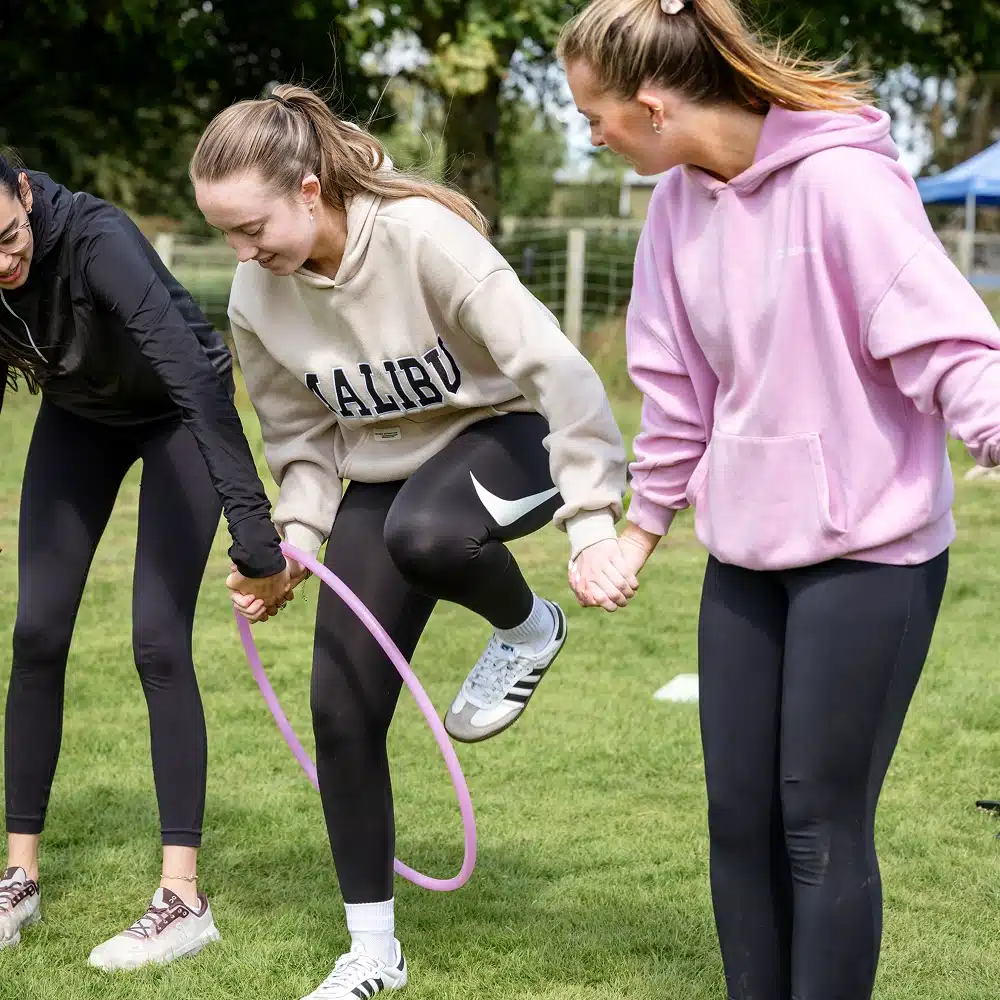 Three young women outdoors hold hands, working together to pass a pink hula hoop along their linked arms. Smiling and having fun on a grassy field under sunny weather, they enjoy one of the Navan team-building activities.