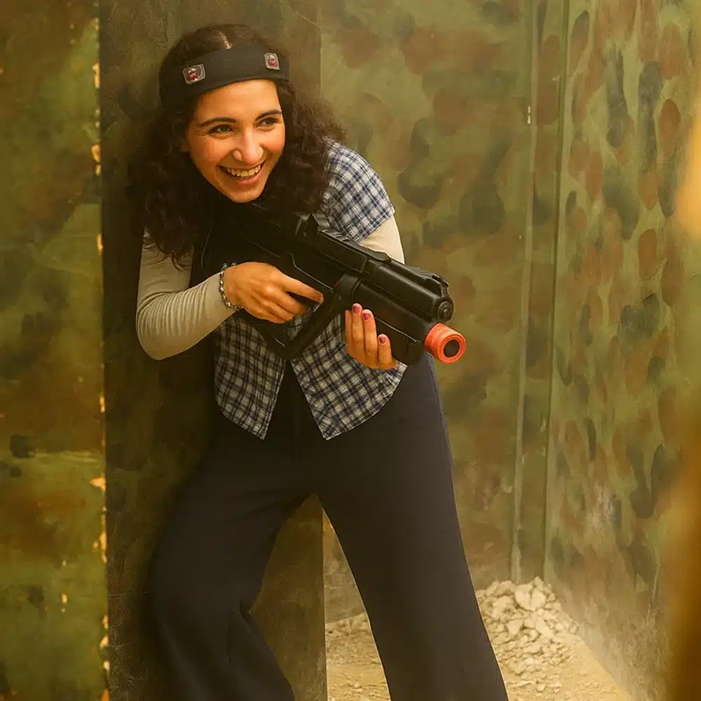 A smiling woman in a headband and checked waistcoat holds a toy gun while leaning against a camouflaged wall, enjoying indoor laser tag as part of Navan team-building activities.