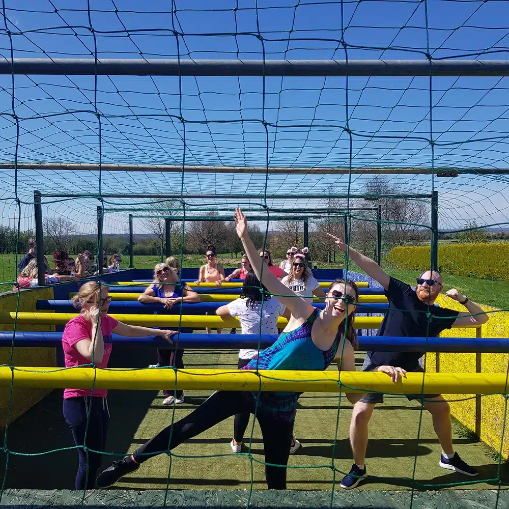 A group of people enjoy human table football outdoors on a sunny day, holding onto horizontal bars and smiling under a netted structure—one of the fun Navan team building activities set amid green fields and yellow barriers.