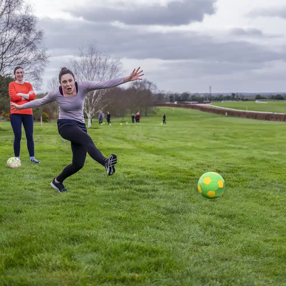 A woman in sports kit kicks a green and yellow football on a grassy pitch, arms flung out for balance, while another woman smiles in the background—perfect for Navan team building activities under a cloudy sky.