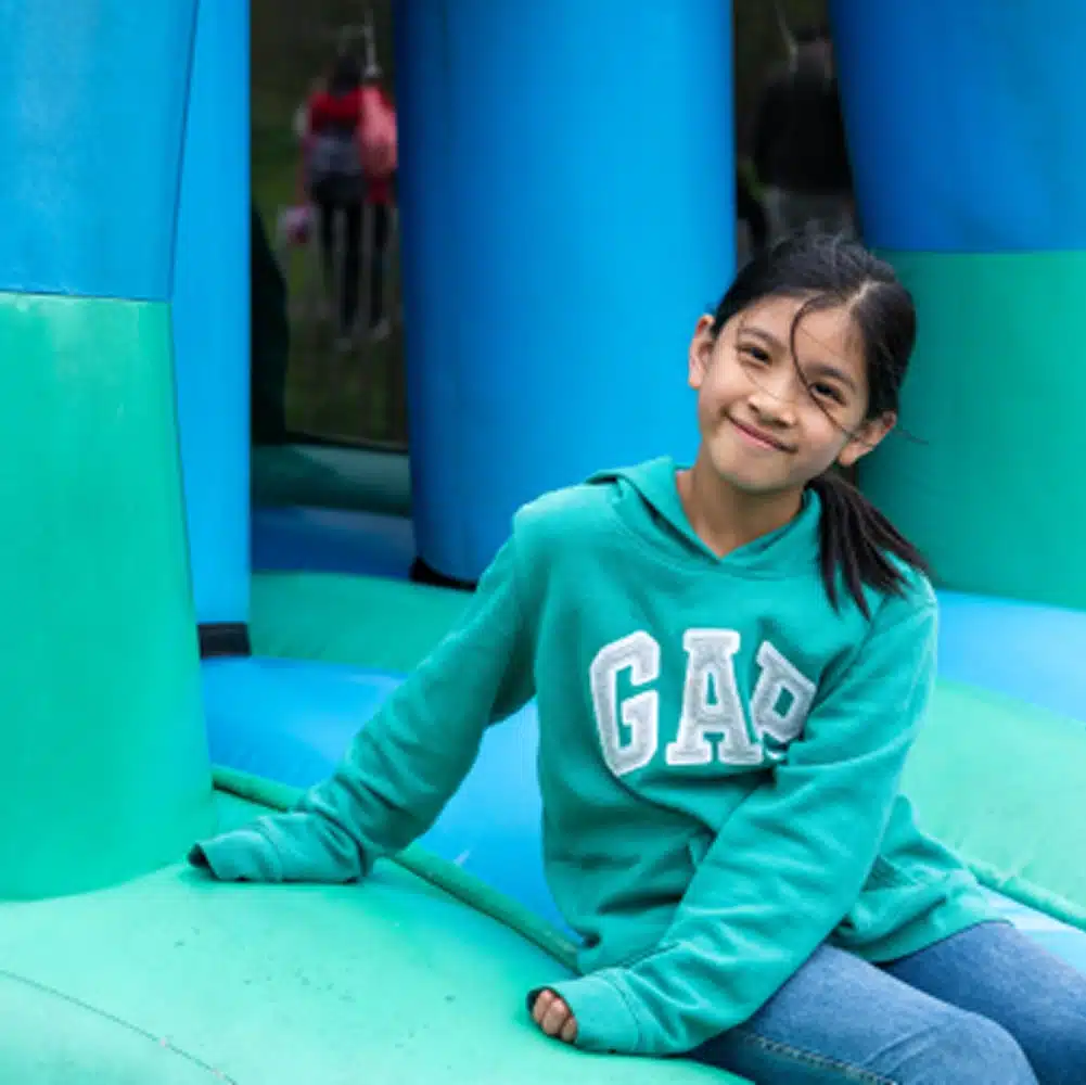 A young girl in a teal GAP hoodie and jeans sits smiling on a blue and green inflatable structure, enjoying the fun at the Navan outdoor inflatables area, possibly during an outdoor event.