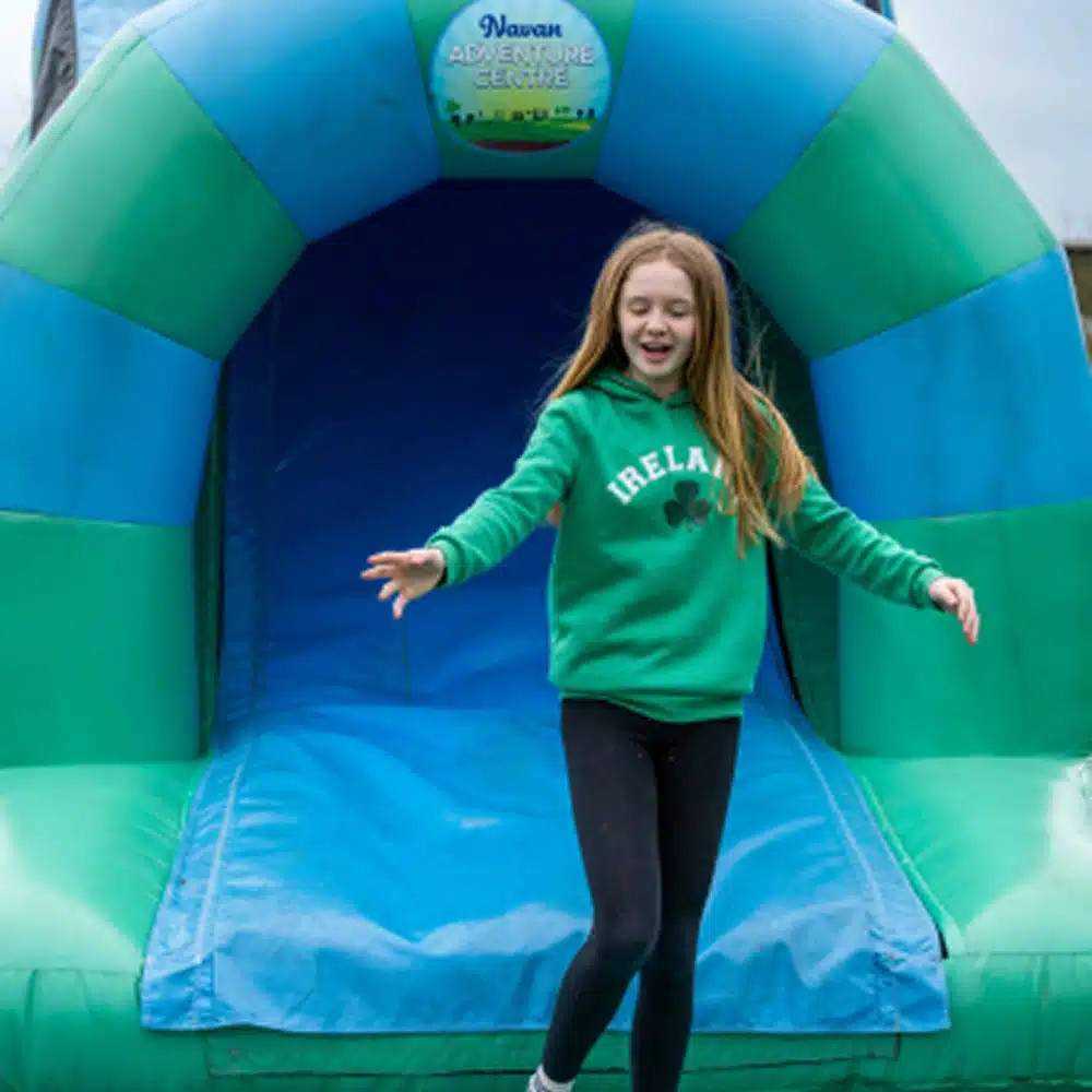 A smiling girl wearing a green "Ireland" sweatshirt and black leggings stands at the entrance of an inflatable slide at the Navan outdoor inflatables Area adventure centre.