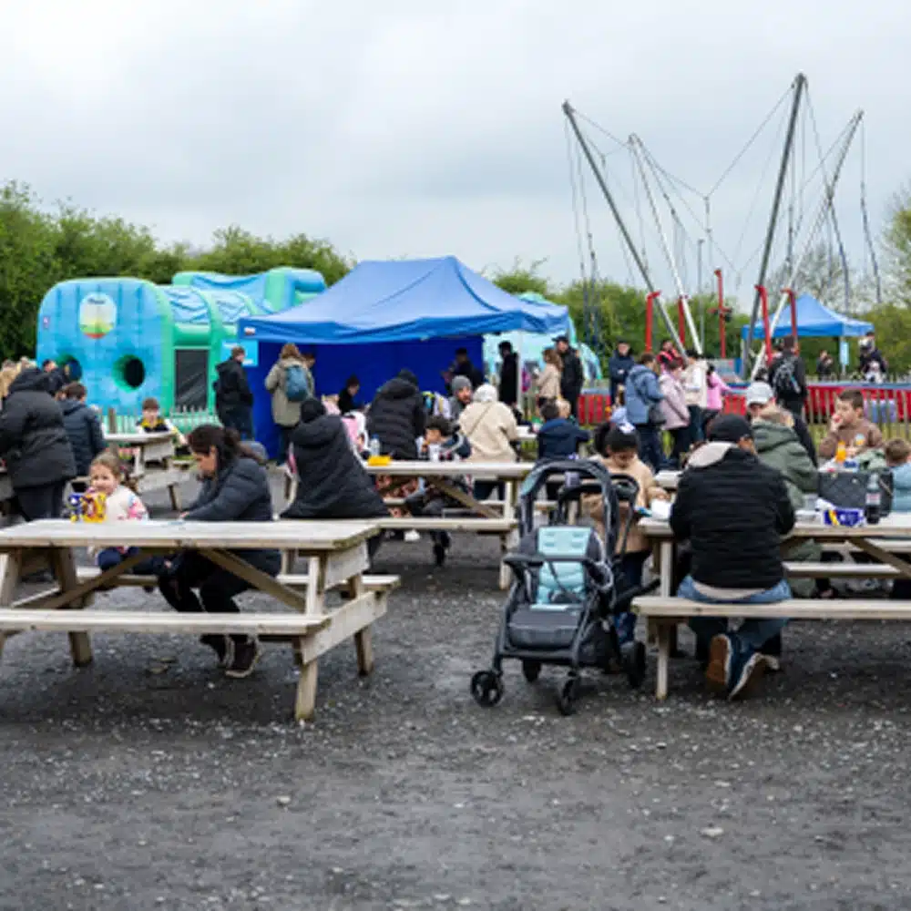 People sit at outdoor picnic tables, some with children and pushchairs, in front of blue marquees and inflatable structures in the Navan outdoor inflatables area. More people stand and socialise nearby at this family-friendly event or fair.
