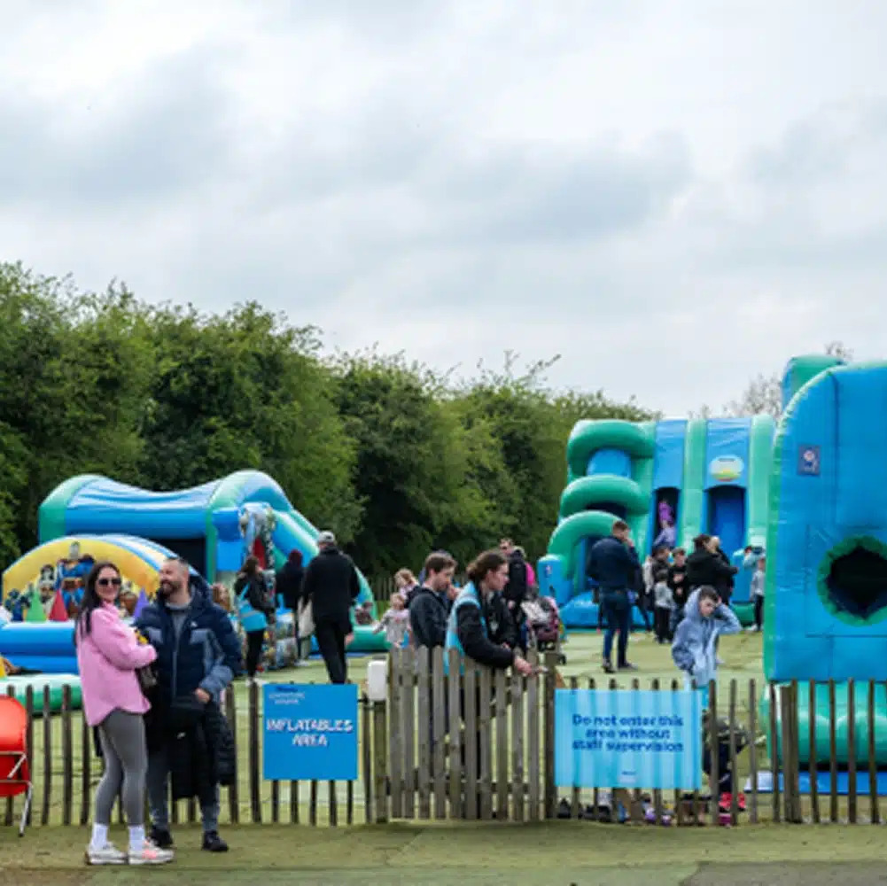 A group of people, including children and adults, gather in the Navan outdoor inflatables area of a park on a cloudy day. Signs and a fence separate the large inflatable play structures from the viewing area.