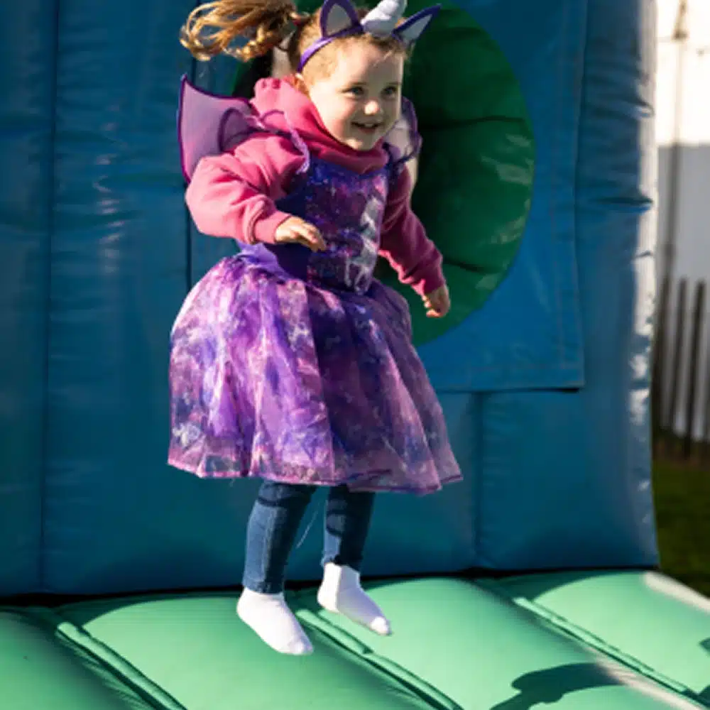 A young child wearing a purple dress, fairy wings, unicorn headband, and white socks is joyfully jumping on a green and blue inflatable bouncy castle in the Navan outdoor inflatables area.