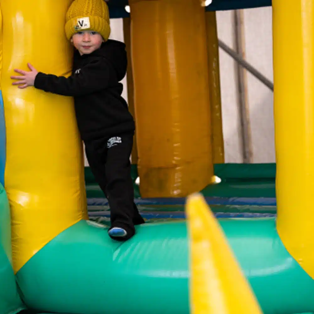 A young child wearing a yellow knitted hat and black outfit hugs the inside wall of a colourful inflatable play structure in the Navan outdoor inflatables area, smiling and balancing with one foot forward.