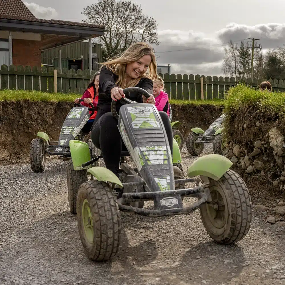 Two smiling women ride green pedal go-karts on a dirt track outdoors, with grass and a wooden fence in the background. It's a fun scene inspired by the Navan junior driving course, as one woman leads and laughs whilst the other follows closely behind.