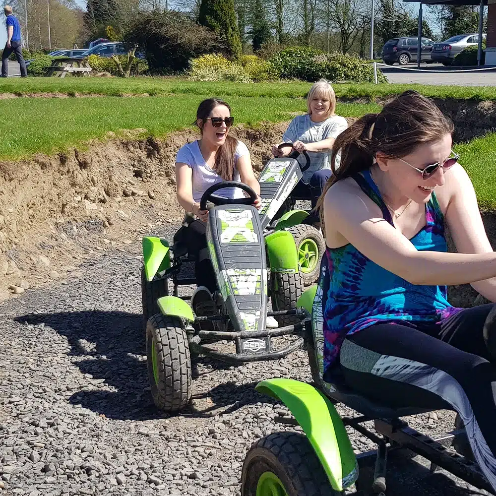 Three women ride green pedal go-karts on a gravel path, smiling and enjoying a sunny day outdoors at the Navan junior driving course, with grass, trees, and parked cars in the background.