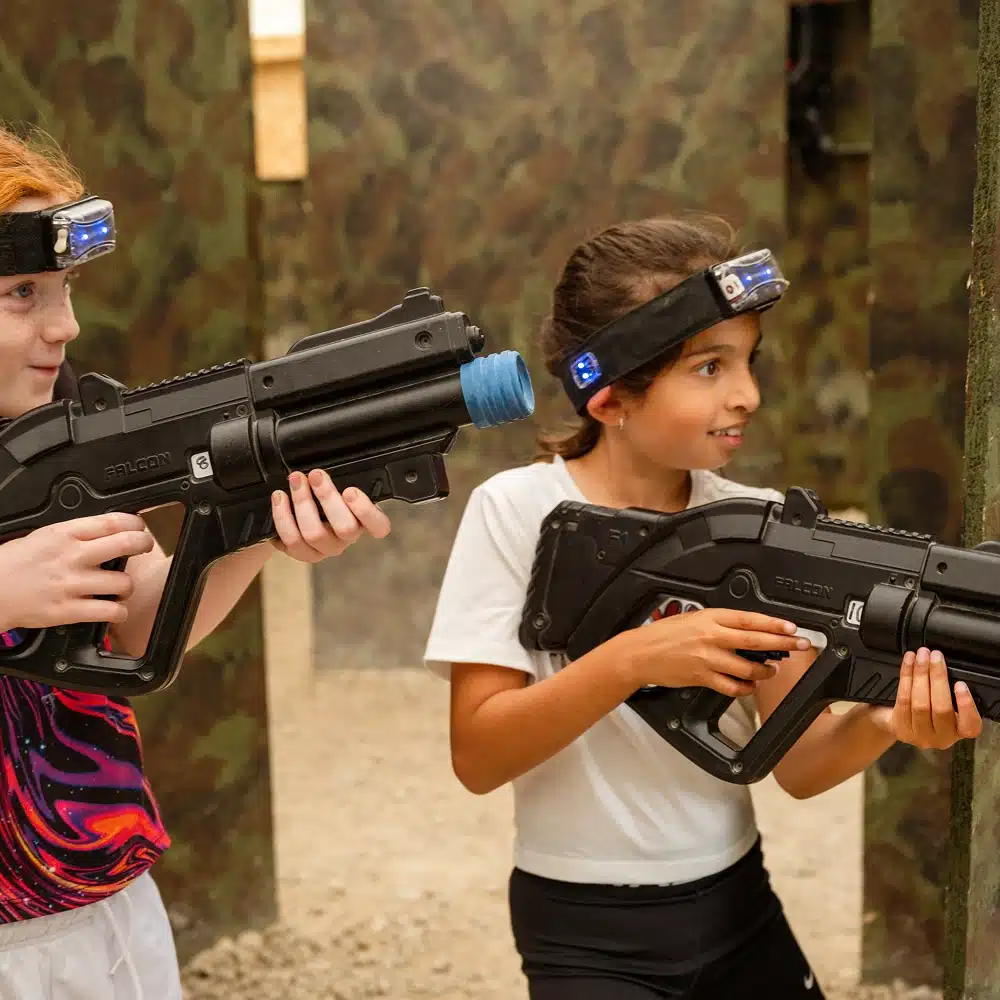 Two children wearing headbands with sensors play a Navan indoor laser tag game, aiming black toy guns and smiling. The background features camouflage-patterned walls and a sandy floor.
