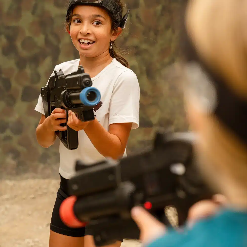 A smiling child wearing a headband and white t-shirt holds a toy laser tag gun, aiming at another player in the foreground. The background features a camouflage-patterned wall and gravel ground at Navan indoor laser tag.