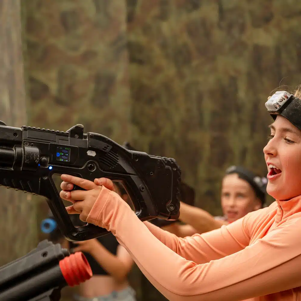 A young person wearing an orange long-sleeved shirt and a headband with a sensor excitedly aims a large black laser tag gun at Navan indoor laser tag. Another player with a similar headband is visible in the background.