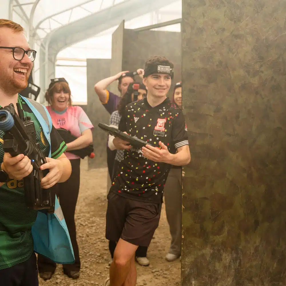 A group of people playing Navan indoor laser tag, smiling and laughing whilst holding laser tag guns, with camouflage barriers and bright lighting in the background.