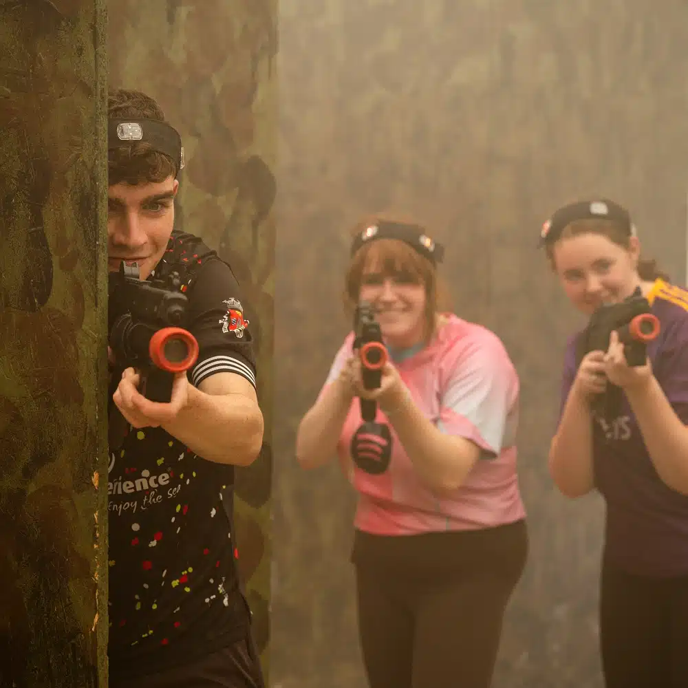 Three people play Navan indoor laser tag in a dim, camouflage-themed arena. One person aims a laser gun from behind a wall whilst two others in colourful shirts aim their guns and smile in the background.