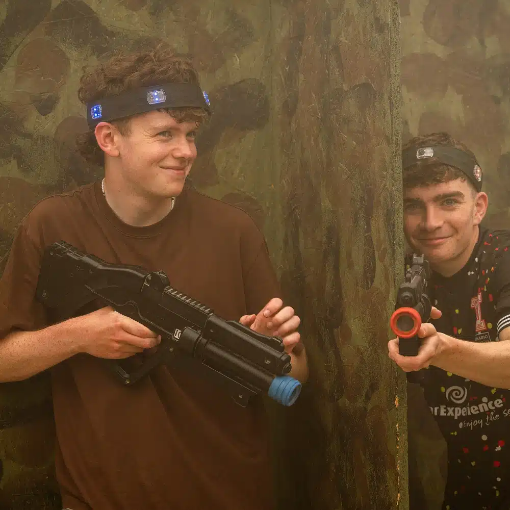 Two young men playing Navan indoor laser tag crouch behind a camouflaged wall. Both are smiling; one holds a laser gun and wears a headband with blue lights, whilst the other aims his laser gun round the corner.