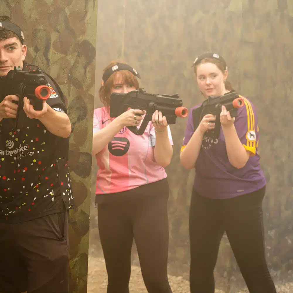 Three people wearing sports shirts and headbands hold black toy laser tag guns, standing in a foggy, camouflaged Navan indoor laser tag arena. One person aims the gun, while the others look towards the camera.