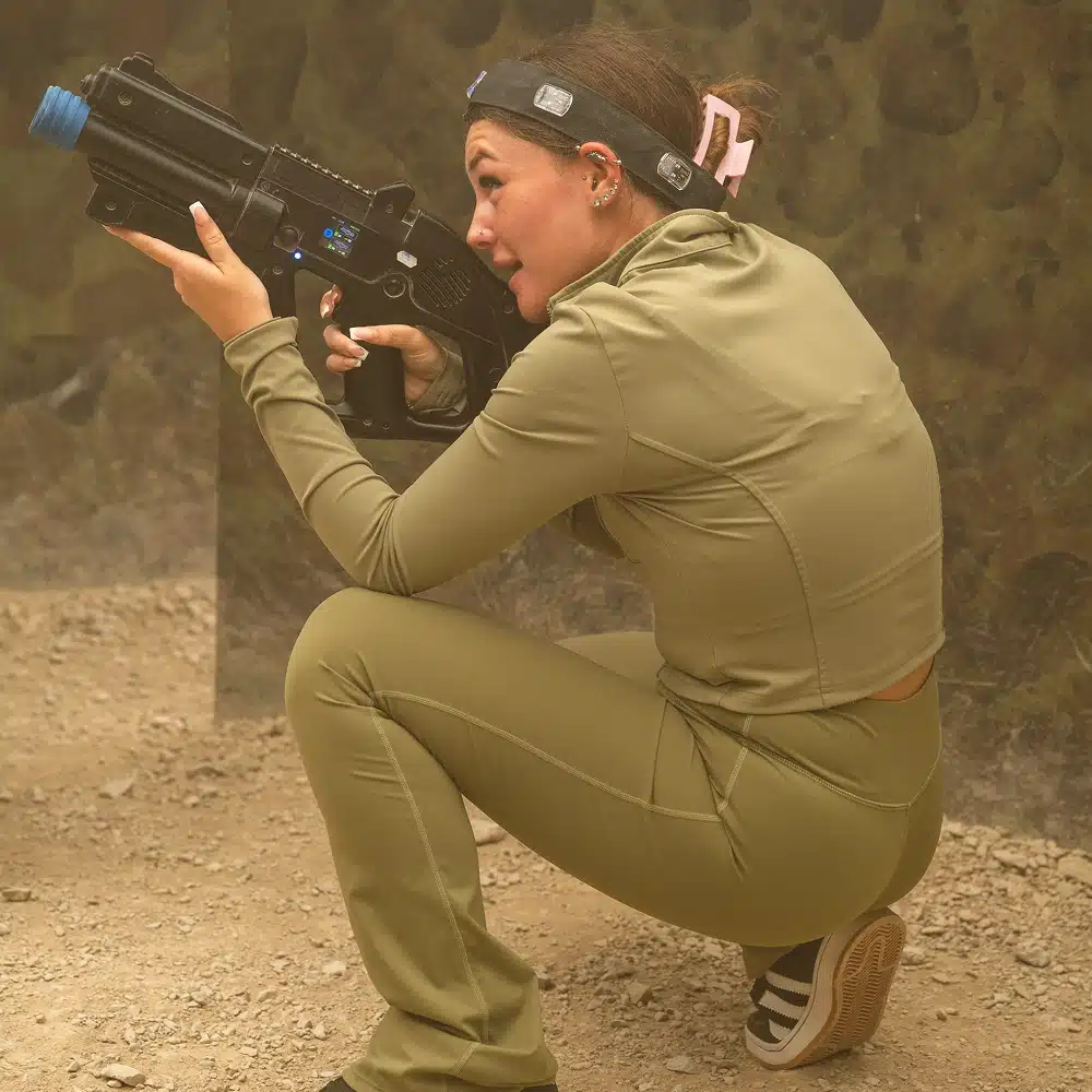 A person wearing an olive-green sports outfit crouches with a laser tag gun in Navan indoor laser tag arena, aiming forward. They have a headband with sensors and a pink bow, surrounded by camouflage walls and sandy ground.