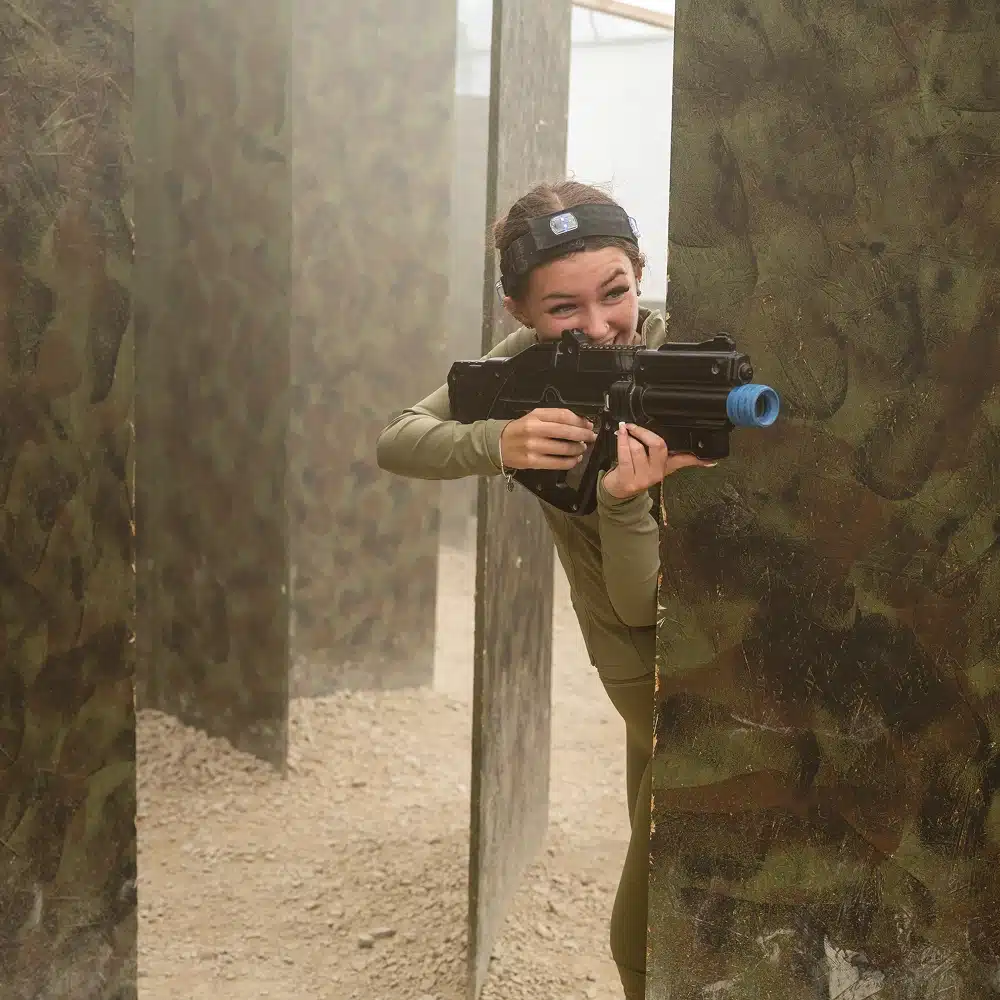 A person wearing a headband and green outfit crouches behind a camouflaged barrier in Navan indoor laser tag arena, smiling and aiming a toy gun with a blue tip.