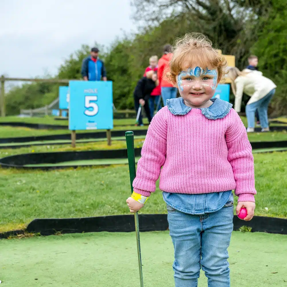 A young child with face paint, wearing a pink jumper and jeans, stands on the Navan mini-golf course holding a club and ball. People and blue "Hole 5" signs are visible in the background.