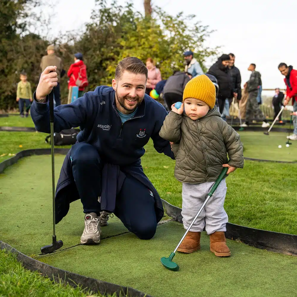 A smiling man kneels beside a young child holding a mini-golf club and golf ball on the grassy Navan mini-golf course, with other people and children enjoying the game in the background.